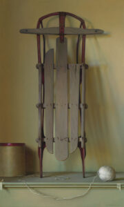 A vintage wooden sled with red metal runners is propped vertically against a plain wall. Beside it, crafted by a talented Duluth MN artist, sits a ball of white string partially unraveled next to a round, tan cardboard box with a red lid.