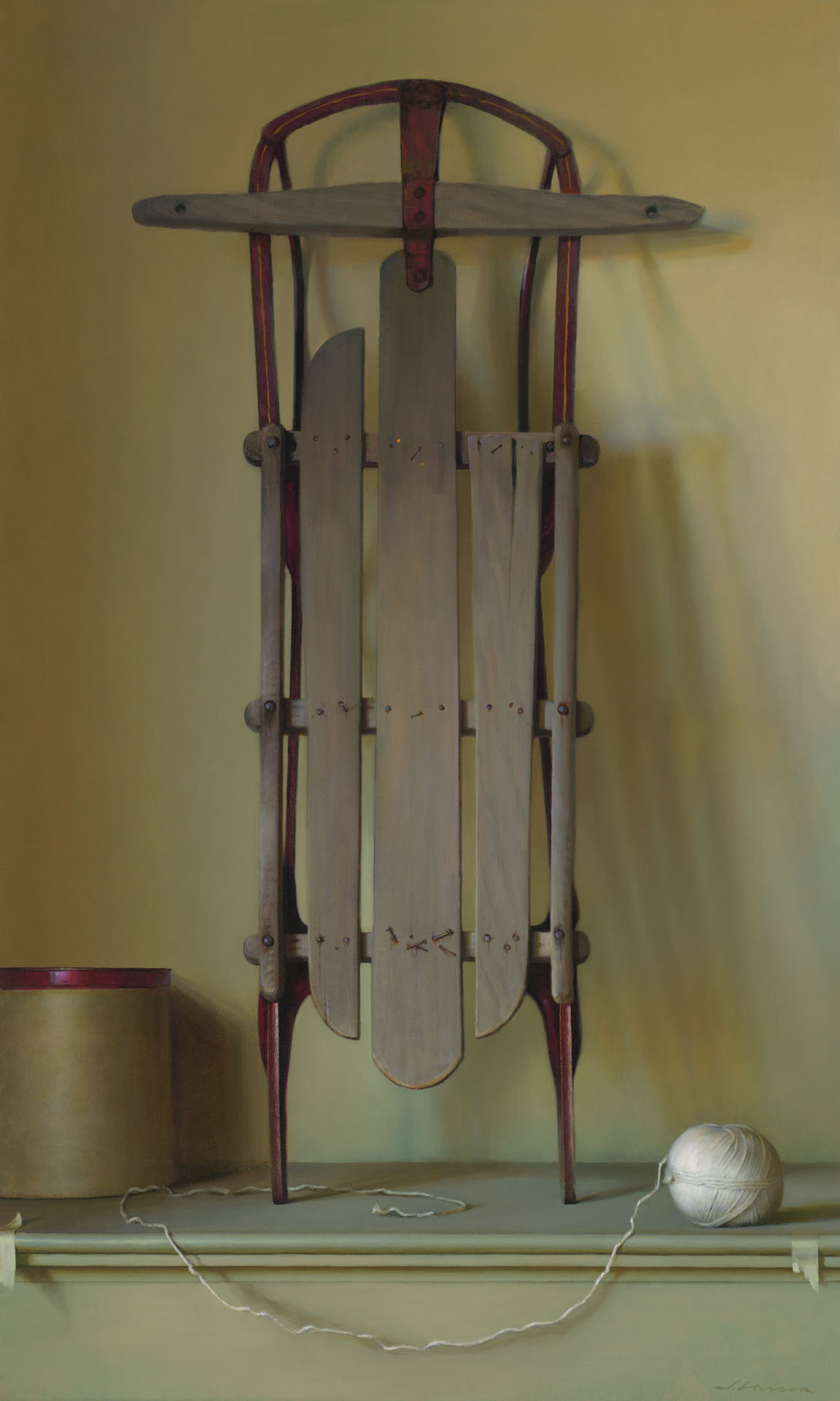 A vintage wooden sled with red metal runners is propped vertically against a plain wall. Beside it, crafted by a talented Duluth MN artist, sits a ball of white string partially unraveled next to a round, tan cardboard box with a red lid.