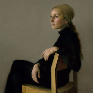 A contemplative Duluth MN artist with long, braided hair sits on a wooden chair. Dressed in black, they wear a pensive expression as their hands rest on the chair's back. The plain background accentuates the artist’s thoughtful pose, highlighting their introspective nature.