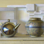 A still life composition featuring a shiny metal teapot, a ceramic vase with blue floral patterns, and geometric white foam shapes arranged on a shelf with a pale yellow background.