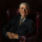 A portrait by a Duluth MN artist captures an older man in a dark leather armchair, dressed in a dark suit with a patterned tie and glasses. He smiles warmly, hands clasped in his lap. The dim background accentuates the subject's presence.