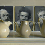 Three beige ceramic pitchers are displayed in front of three abstract black-and-white portraits pinned to a wall. The arrangement creates a juxtaposition of shapes and styles, blending realism with abstract art.