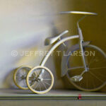A vintage tricycle is depicted with a muted color palette. Its old-fashioned design is accented by soft lighting. A small red toy lies on the floor beneath it, contrasting with the tricycle's subdued tones.
