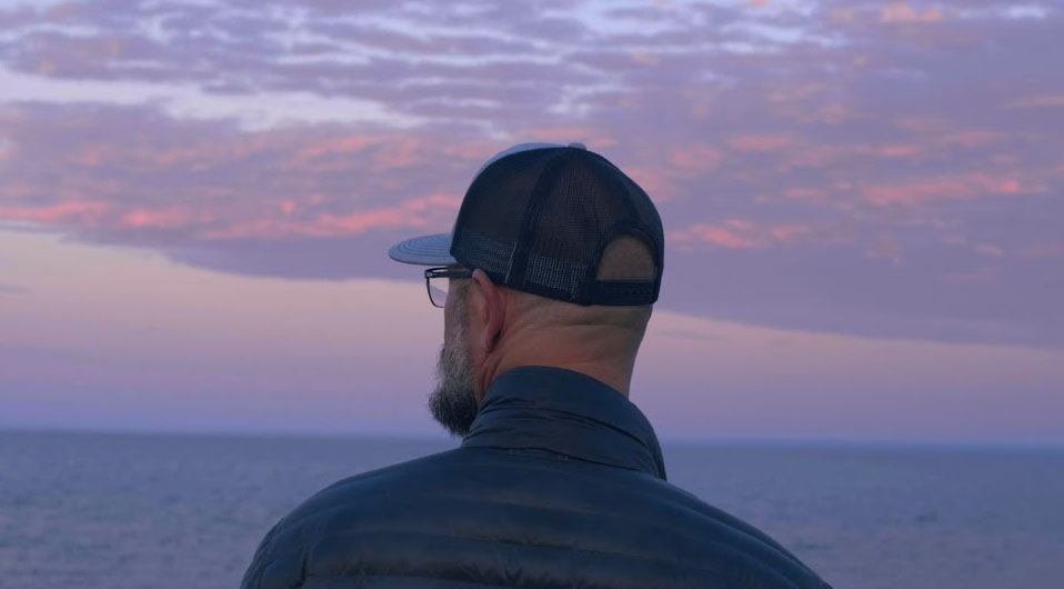 A person wearing a baseball cap and glasses stands facing the ocean, gazing at the sunset. The sky is filled with pink and purple clouds, blending into the horizon over calm water. The person wears a dark jacket, and the scene feels peaceful.