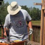 A person wearing a wide-brimmed hat and a gray T-shirt is painting outdoors. They are standing near an easel with a Greek flag draped over it, surrounded by art supplies. Trees and a grassy field are visible in the background.