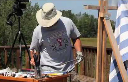 A person wearing a wide-brimmed hat and a gray T-shirt is painting outdoors. They are standing near an easel with a Greek flag draped over it, surrounded by art supplies. Trees and a grassy field are visible in the background.