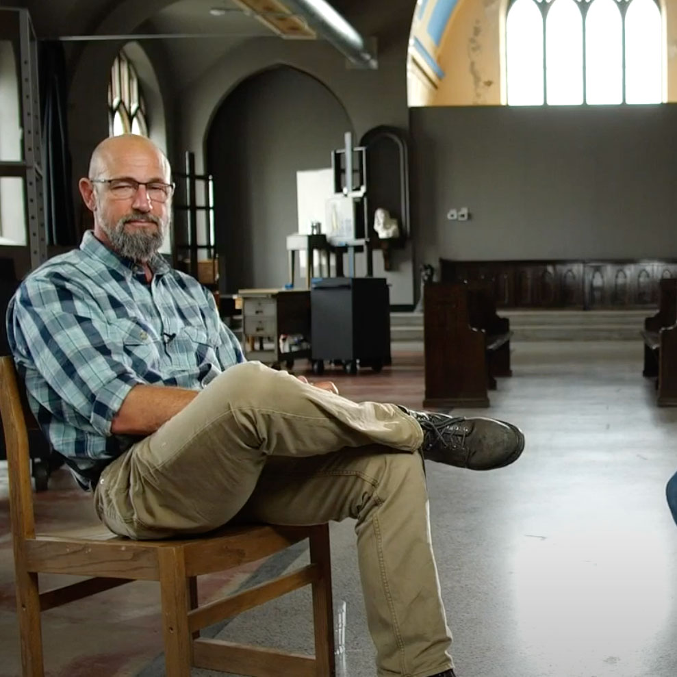 A man with a beard and glasses sits on a wooden chair with legs crossed in a spacious room with high ceilings and arched windows. Natural light streams in, illuminating the room decorated with various pieces of equipment.