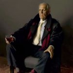 An older man wearing a dark coat and a white tie sits in an armchair holding a glass of red drink, looking serious in a dimly lit setting.